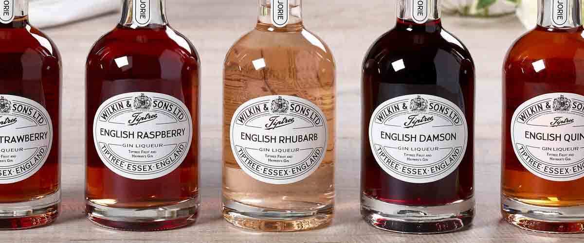 Tiptree-Wilkin-&-Sons-English-gin-liqueurs-Strawberry,-Raspberry,-Rhubarb,-Damson-and-Quince-arranged-on-a-wooden-table-with-fresh-fruit,-flowers-and-a-vintage-Tiptree-jam-tin-in-the-background.