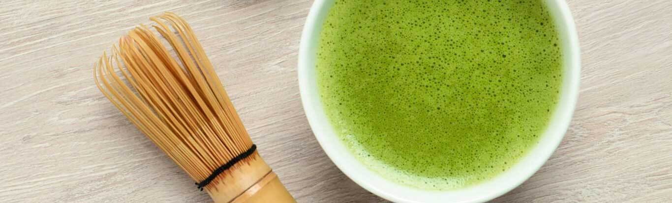 A bowl of freshly whisked vibrant green matcha tea alongside a traditional bamboo whisk on a light wooden surface.